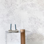 A modernist end table stands in front of a wall papered in a painterly palm tree print in light pink on a cream field.