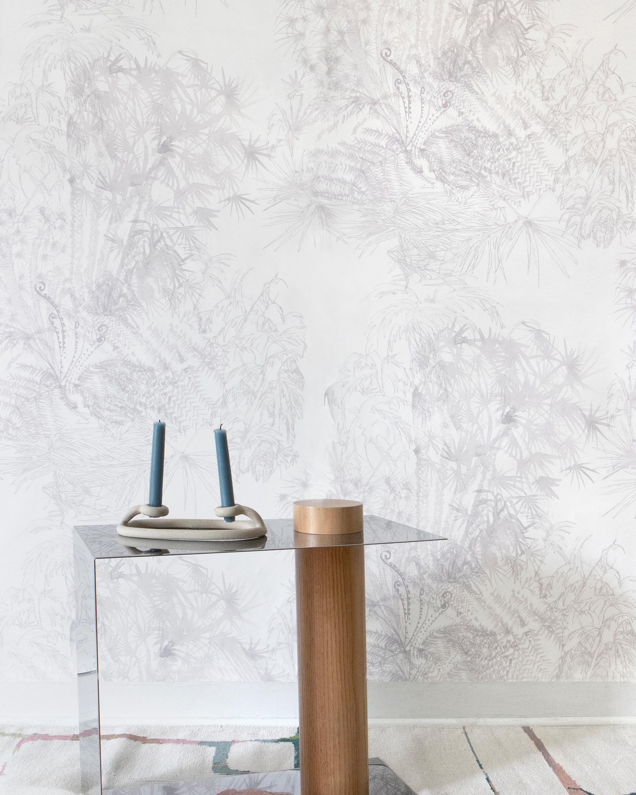 A modernist end table stands in front of a wall papered in a painterly palm tree print in light pink on a cream field.