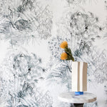 A modernist end table and flowers stands in front of a wall papered in a painterly palm tree print in gray on a cream field.