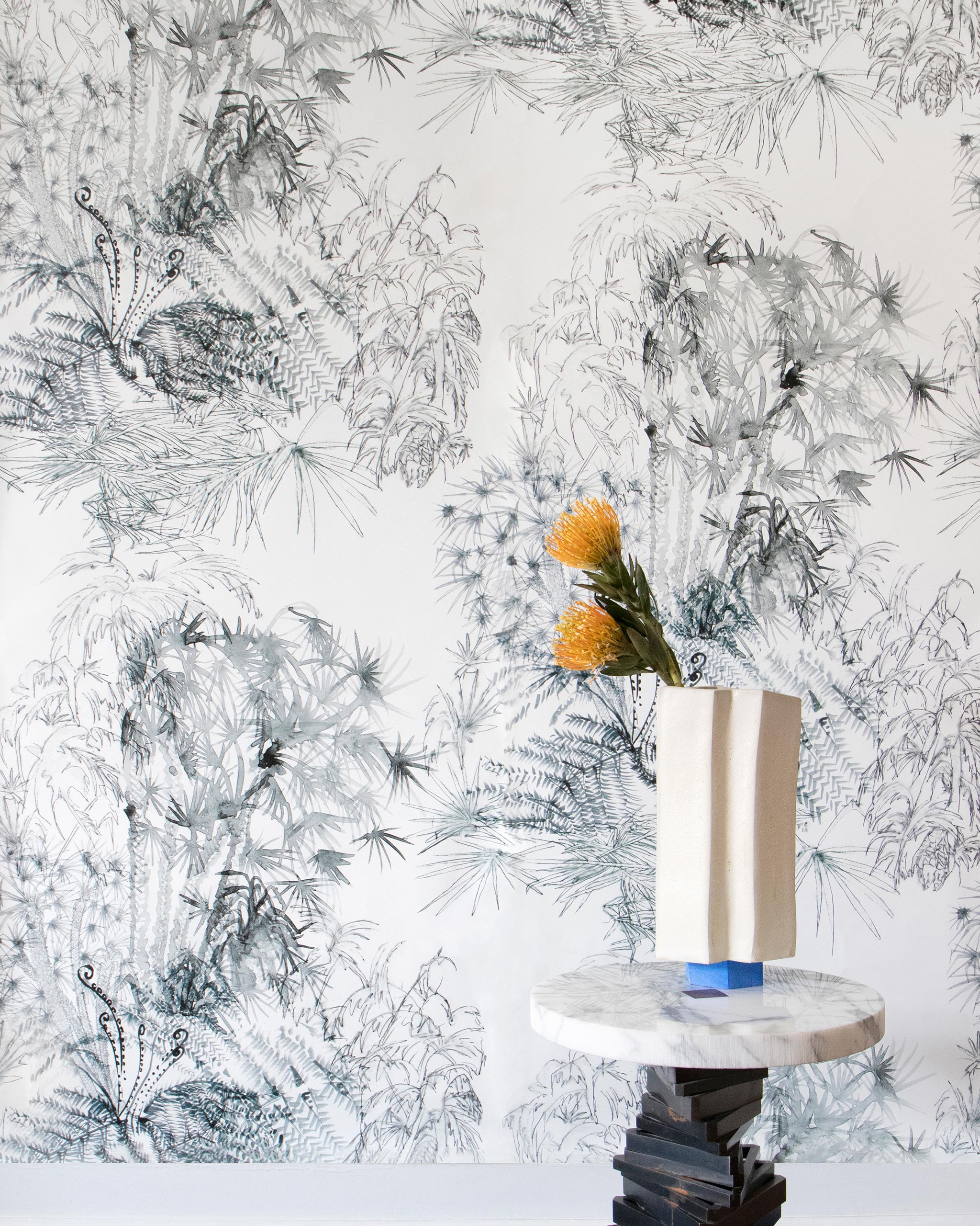 A modernist end table and flowers stands in front of a wall papered in a painterly palm tree print in gray on a cream field.