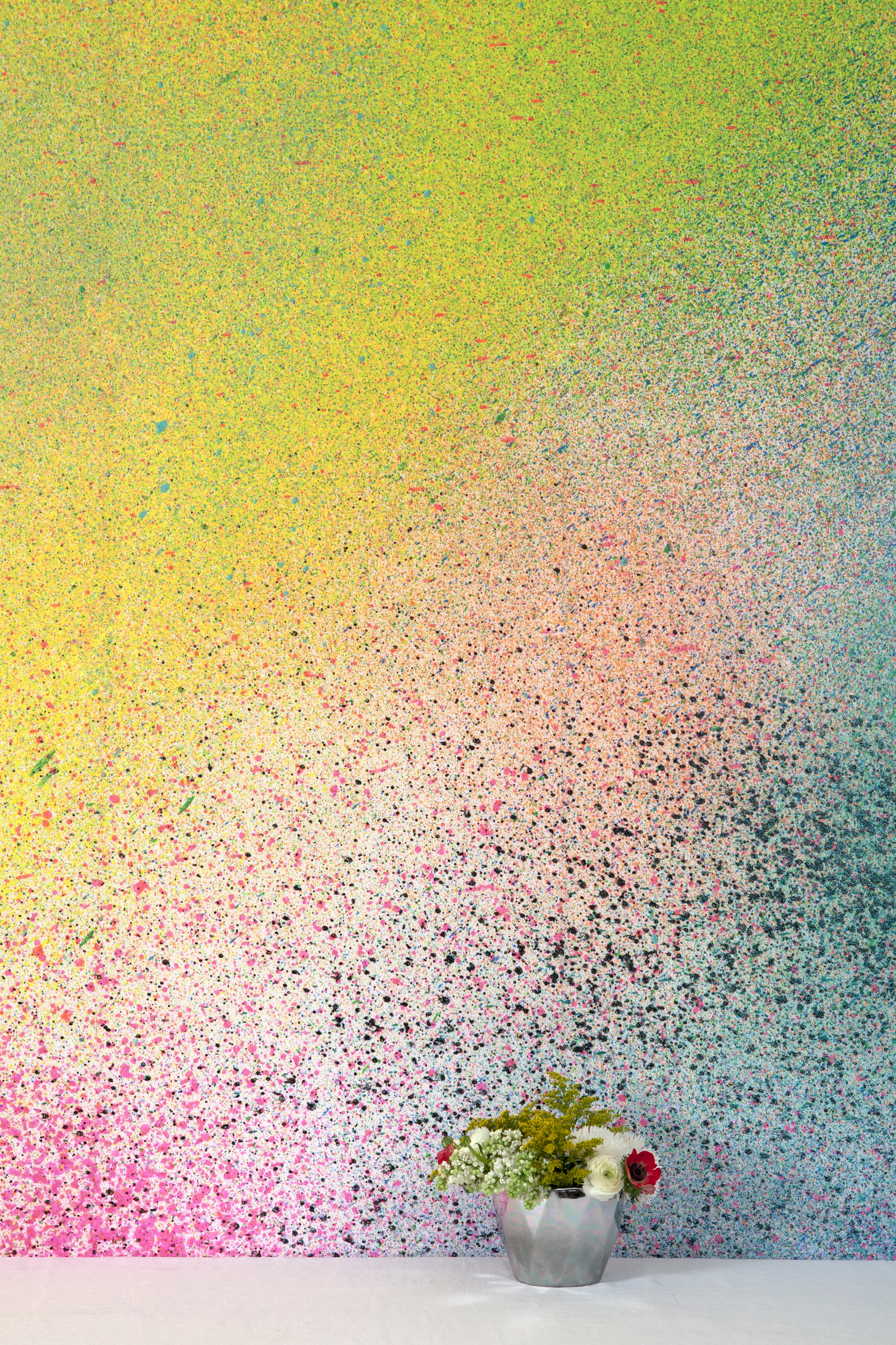 A vase of flowers stands in front of a wall covered in an abstract ombré paint splatter print in pink, yellow, blue and green.