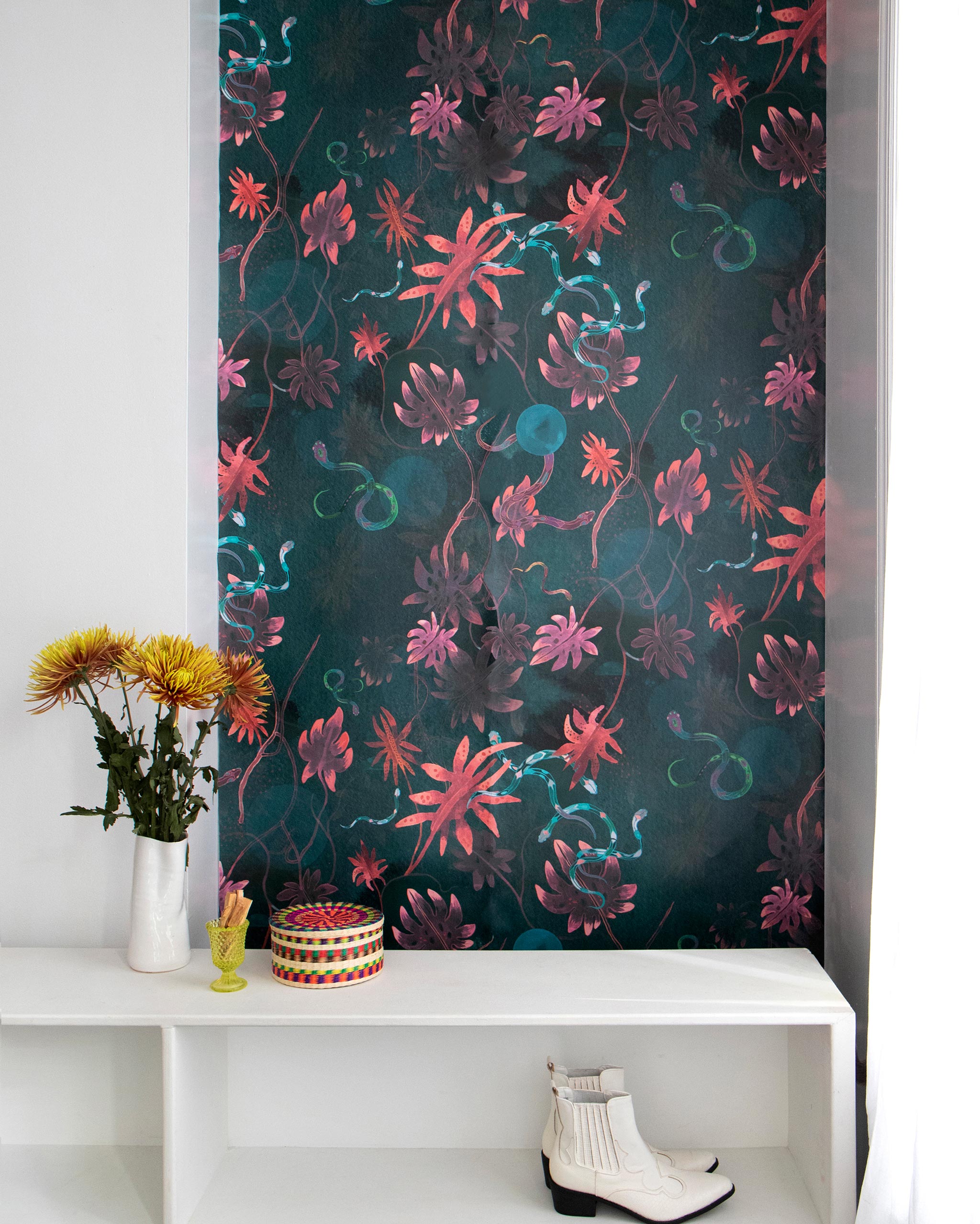 A cluttered end table stands in front of a wall papered in a playful leaf and snake print in pink, green and dark turquoise.