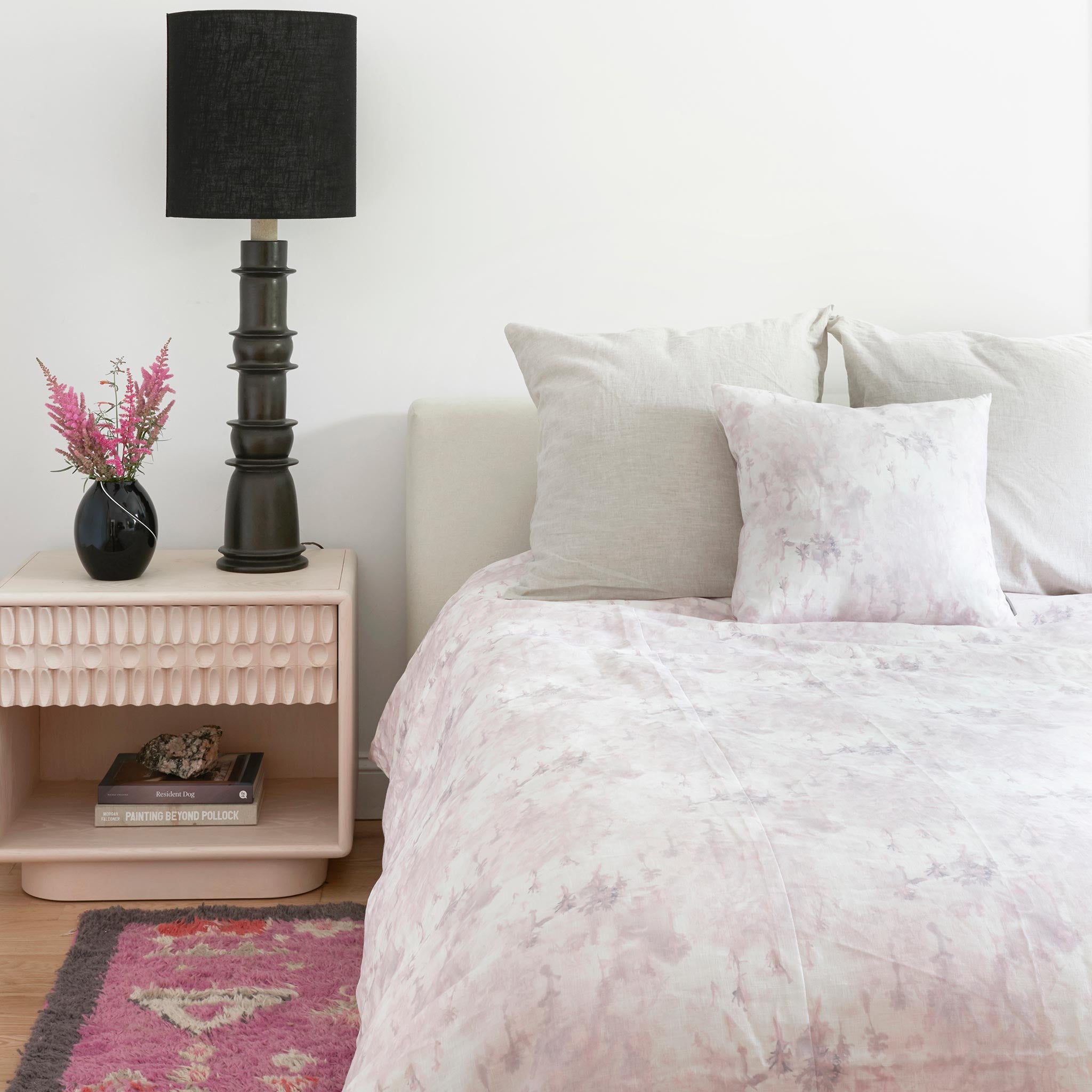 A cozy bedroom tableau with a bed made up in a comforter in a painterly tree print in light pink and purple on a cream field.