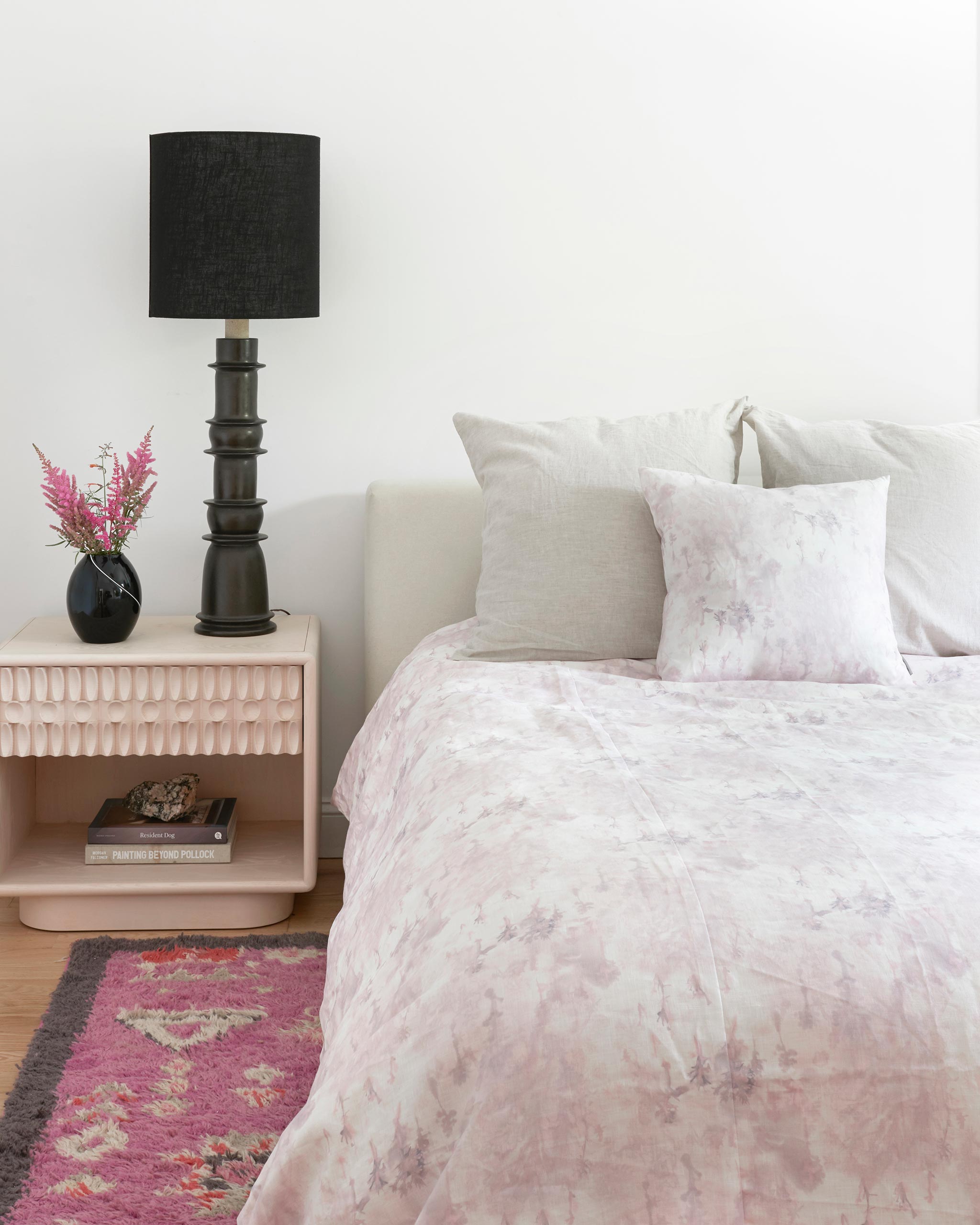 A cozy bedroom tableau with a bed made up in a comforter in a painterly tree print in light pink and purple on a cream field.