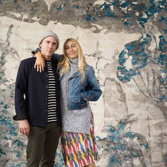 A beachy blonde-haired woman stands with her arm draped around a tall man in front of a large-scale watercolor floral print.