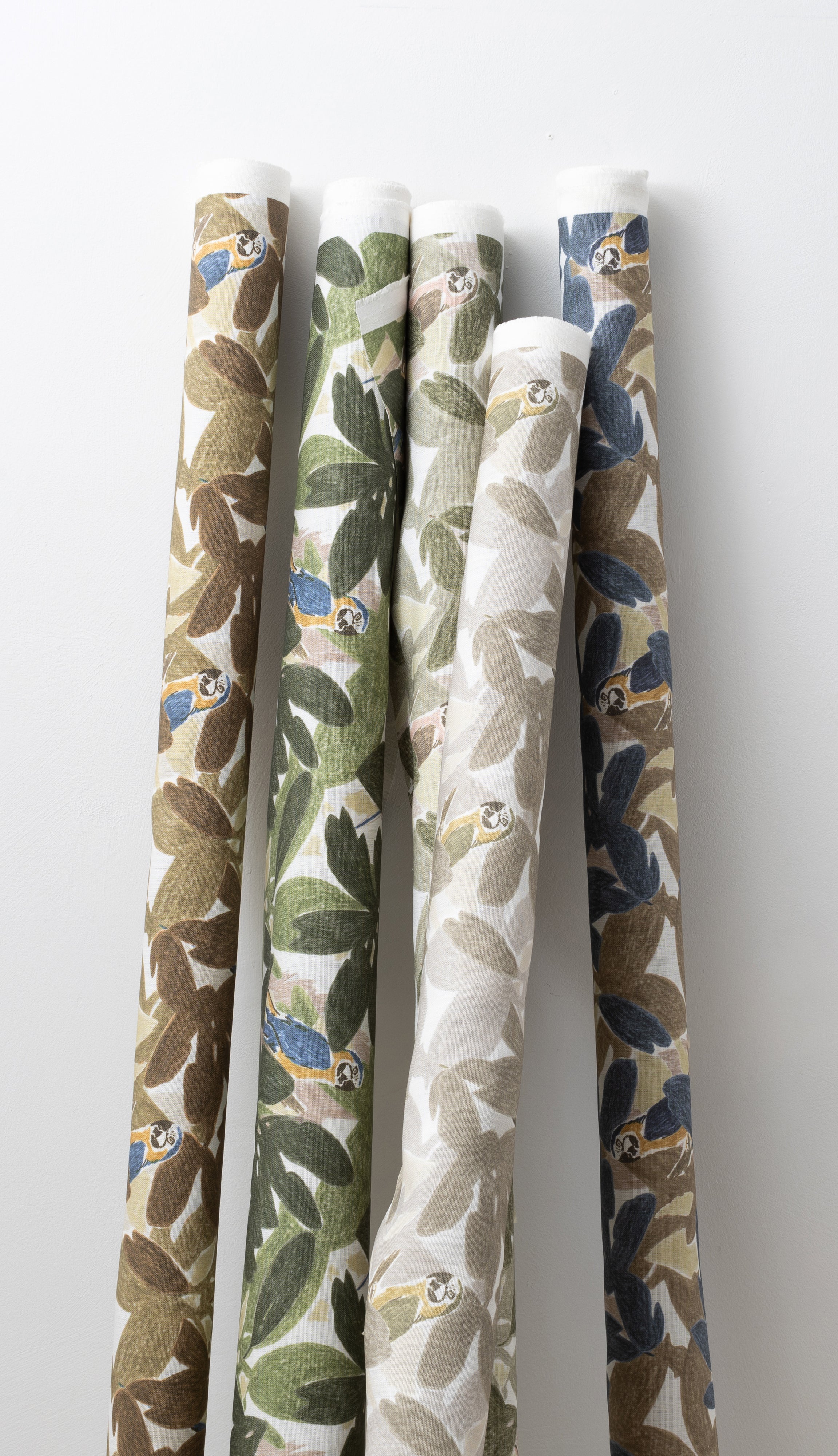 A group of fabric rolls leaned against a wall, all in different colorways of the same botanical and parrot print.