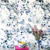 A cluttered end table stands in front of a wall papered in an abstract paint blotch print in black, gray, blue, green and white.