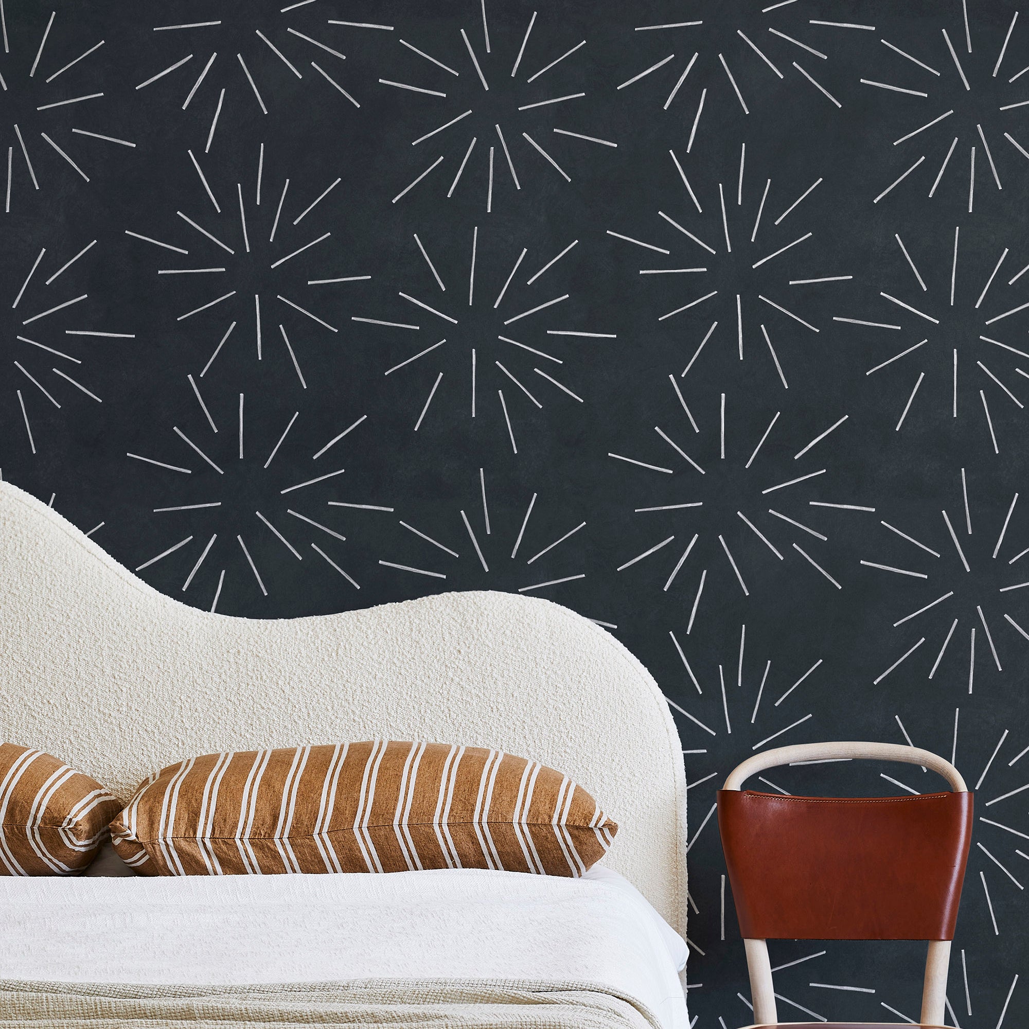 A modernist bed, hanging lamp and chair stand in front of a wall papered in a playful firework print in white and charcoal.