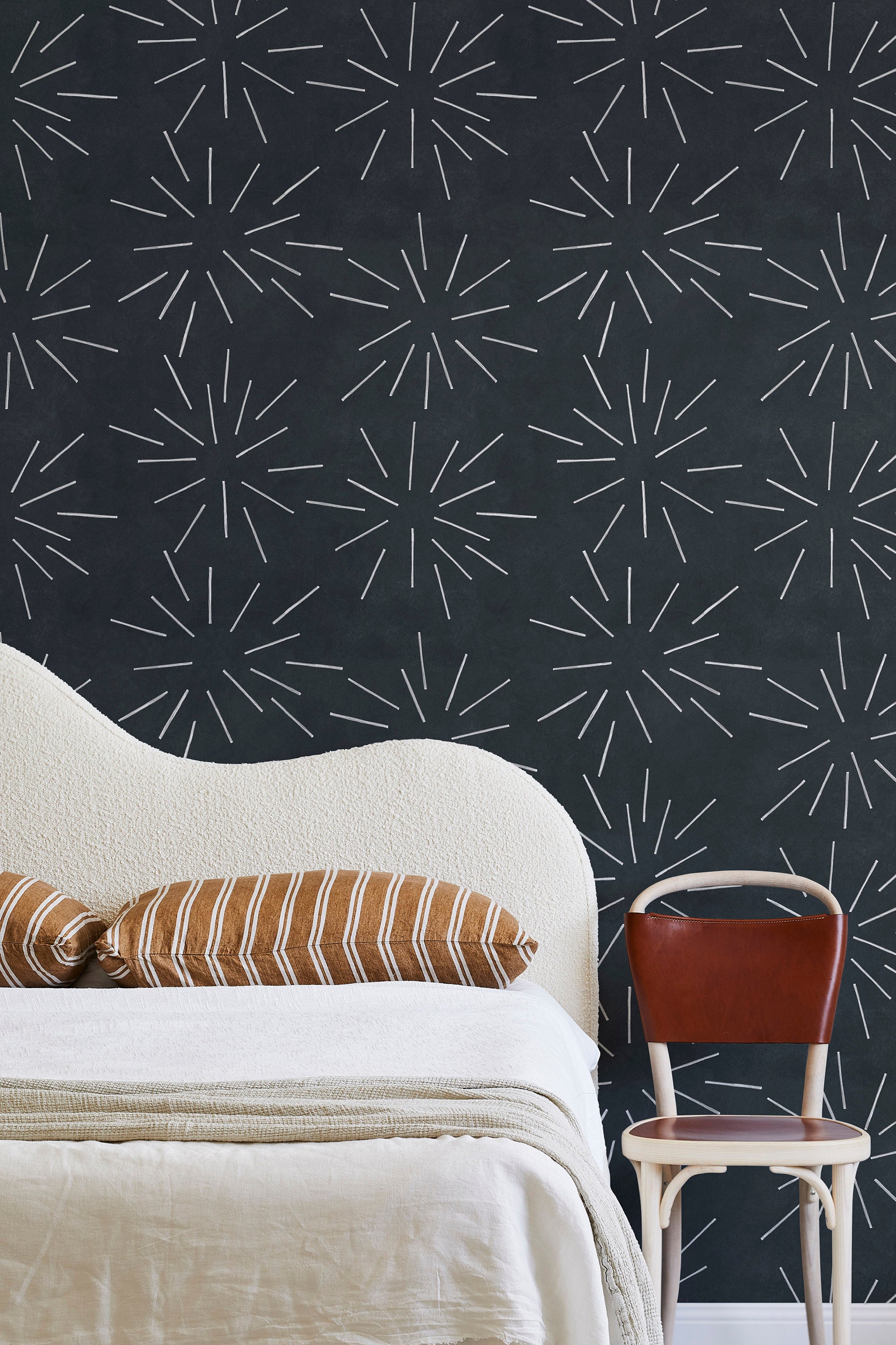 A modernist bed, hanging lamp and chair stand in front of a wall papered in a playful firework print in white and charcoal.