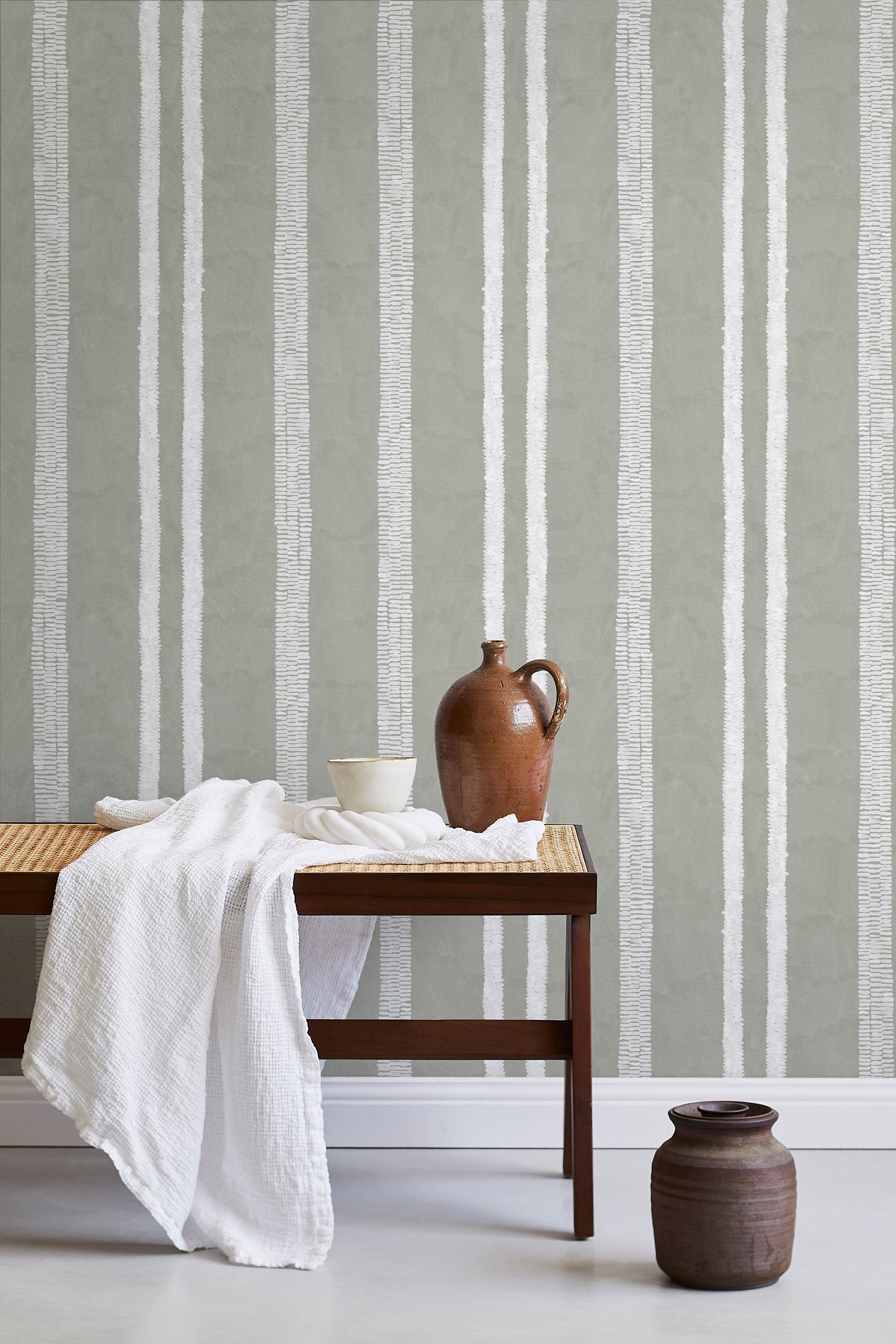 A bench with a towel and clay vases stands in front of a wall papered in a textural stripe pattern in white and greige.