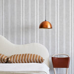 A modernist bed, hanging lamp and chair stand in front of a wall papered in a textural stripe pattern in white and light gray.
