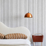 A modernist bed, hanging lamp and chair stand in front of a wall papered in a textural stripe pattern in white and light gray.