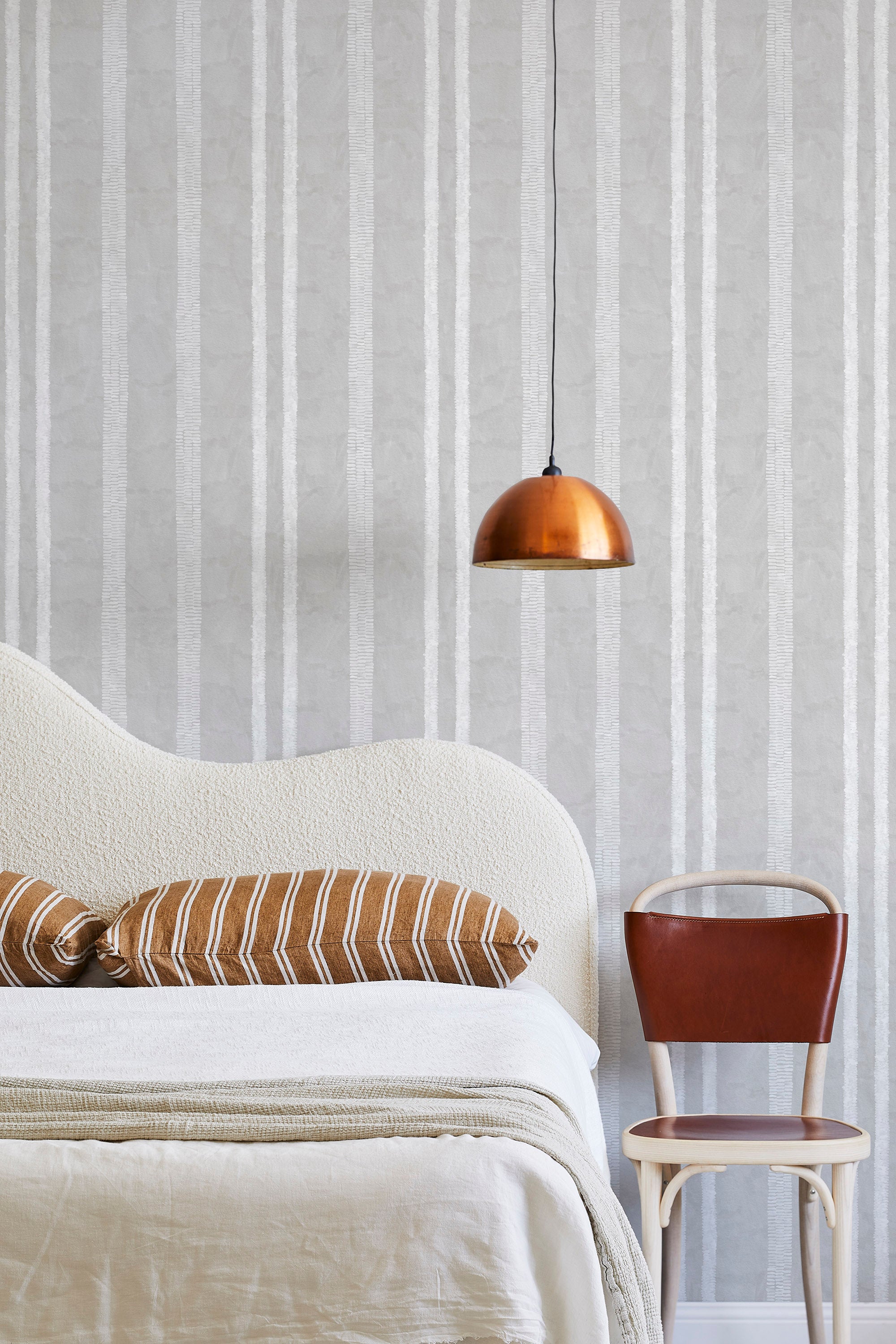 A modernist bed, hanging lamp and chair stand in front of a wall papered in a textural stripe pattern in white and light gray.