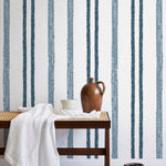 A bench with a towel and clay vases stands in front of a wall papered in a textural stripe pattern in navy and white.