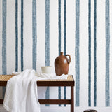 A bench with a towel and clay vases stands in front of a wall papered in a textural stripe pattern in navy and white.