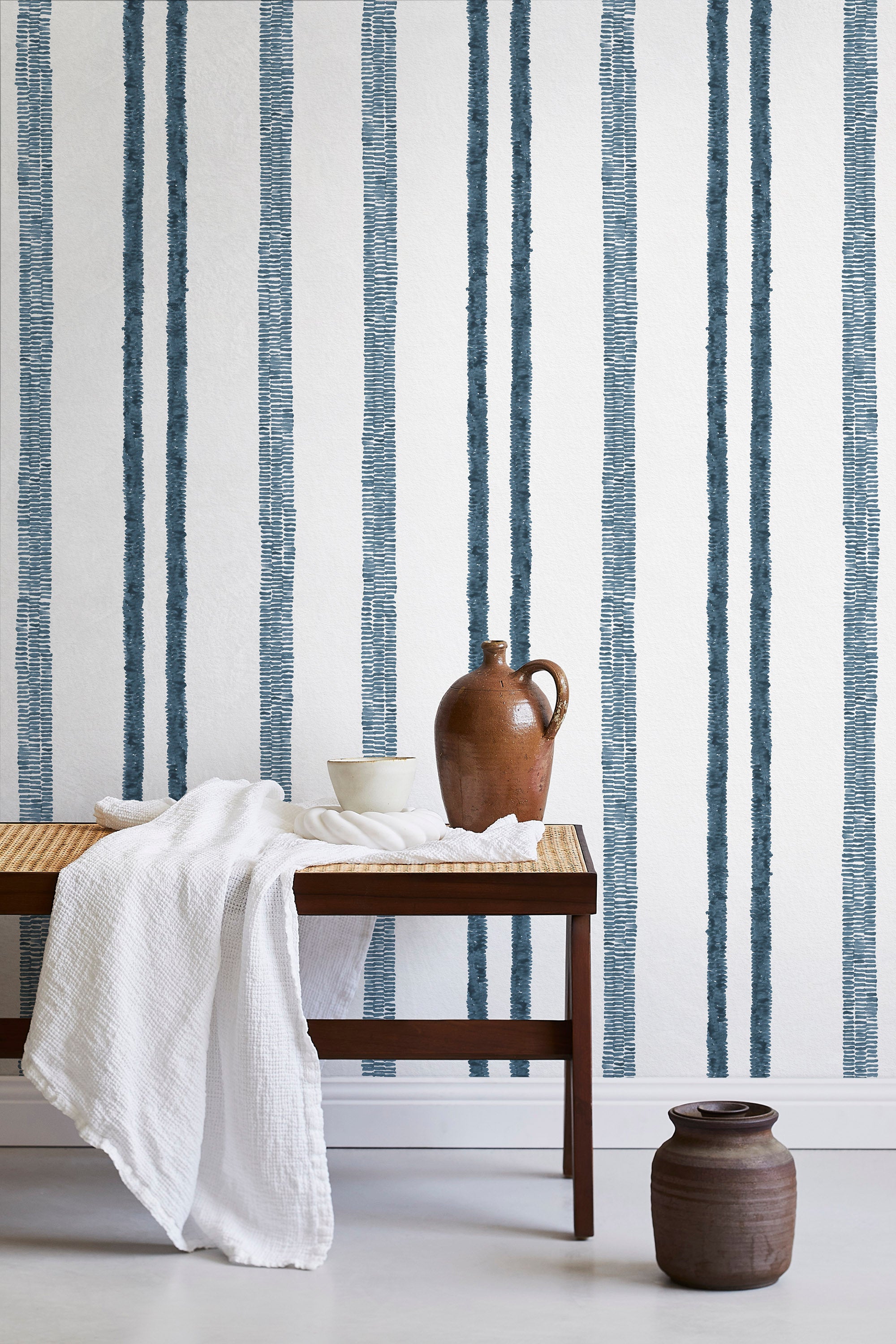 A bench with a towel and clay vases stands in front of a wall papered in a textural stripe pattern in navy and white.