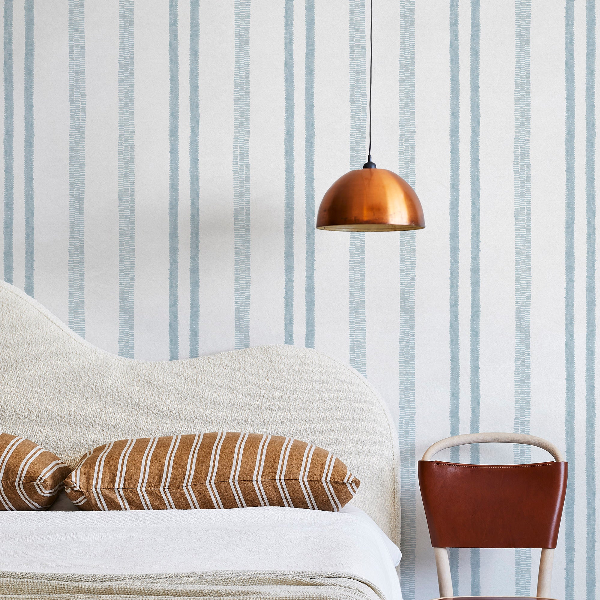 A modernist bed, hanging lamp and chair stand in front of a wall papered in a textural stripe pattern in light blue and white.