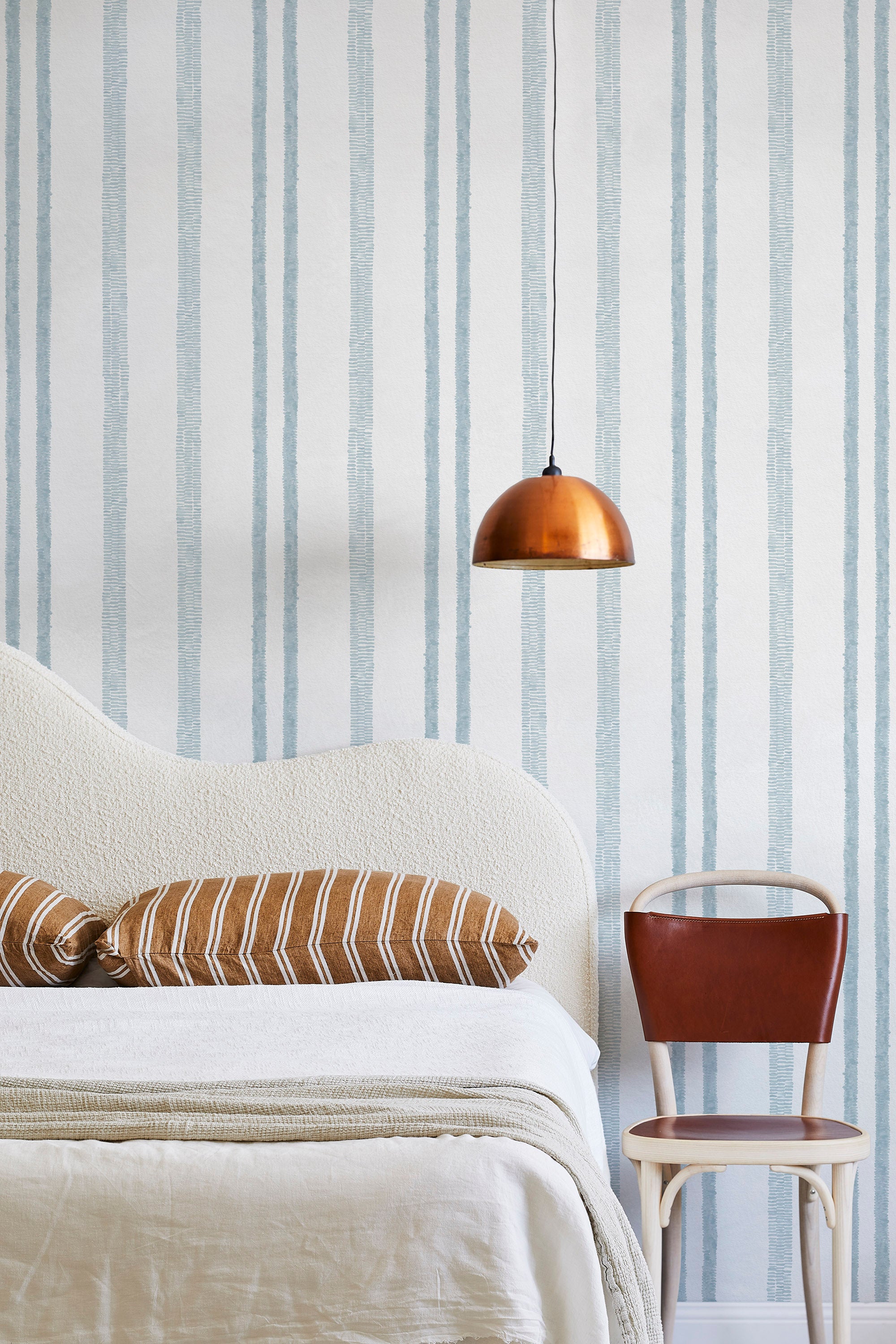 A modernist bed, hanging lamp and chair stand in front of a wall papered in a textural stripe pattern in light blue and white.