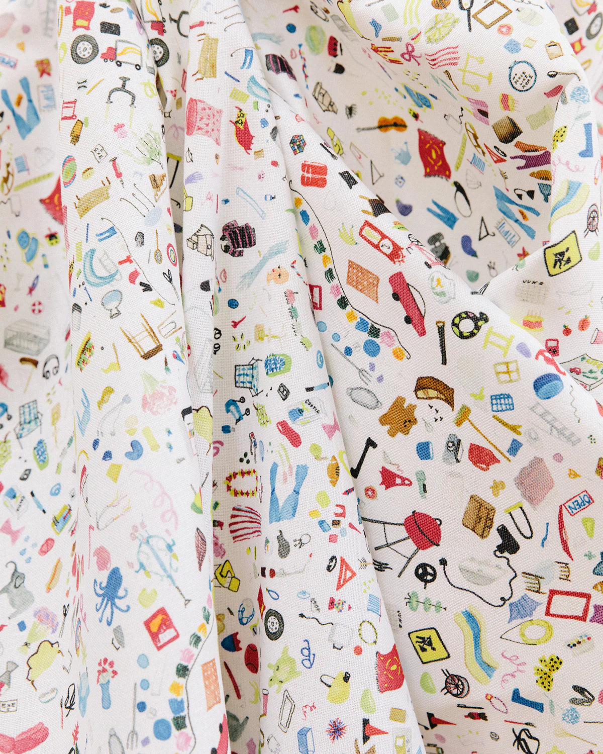 Draped fabric yardage in a playful illustrated "stuff" print in multicolor pastels on a white field.