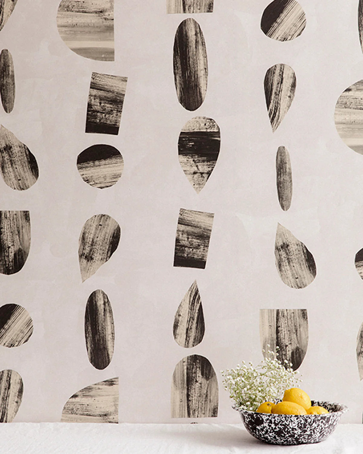 A bowl of fruit stands in front of a wall papered in an abstract curvolinear pattern in black, white and gray.