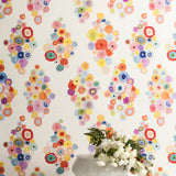 A vase of flowers stands in front of a wall papered in a painterly floral print in multicolor pastels on a white field.