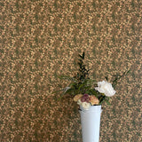 A vase of flowers stands in front of a wall papered in a dense small-scale floral print in shades of brown, tan and green.