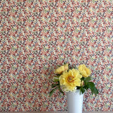 A vase of flowers stands in front of a wall papered in a dense small-scale floral print in shades of coral, red, green and blue.