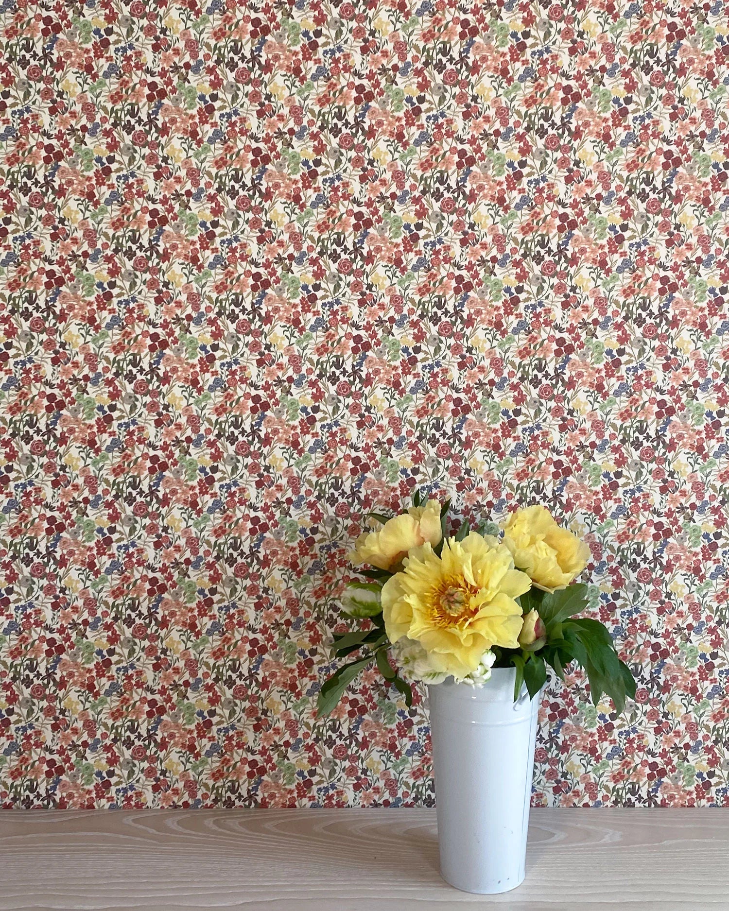 A vase of flowers stands in front of a wall papered in a dense small-scale floral print in shades of coral, red, green and blue.