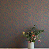 A vase of flowers stands in front of a wall papered in a dense small-scale floral print in shades of purple, gold and navy.