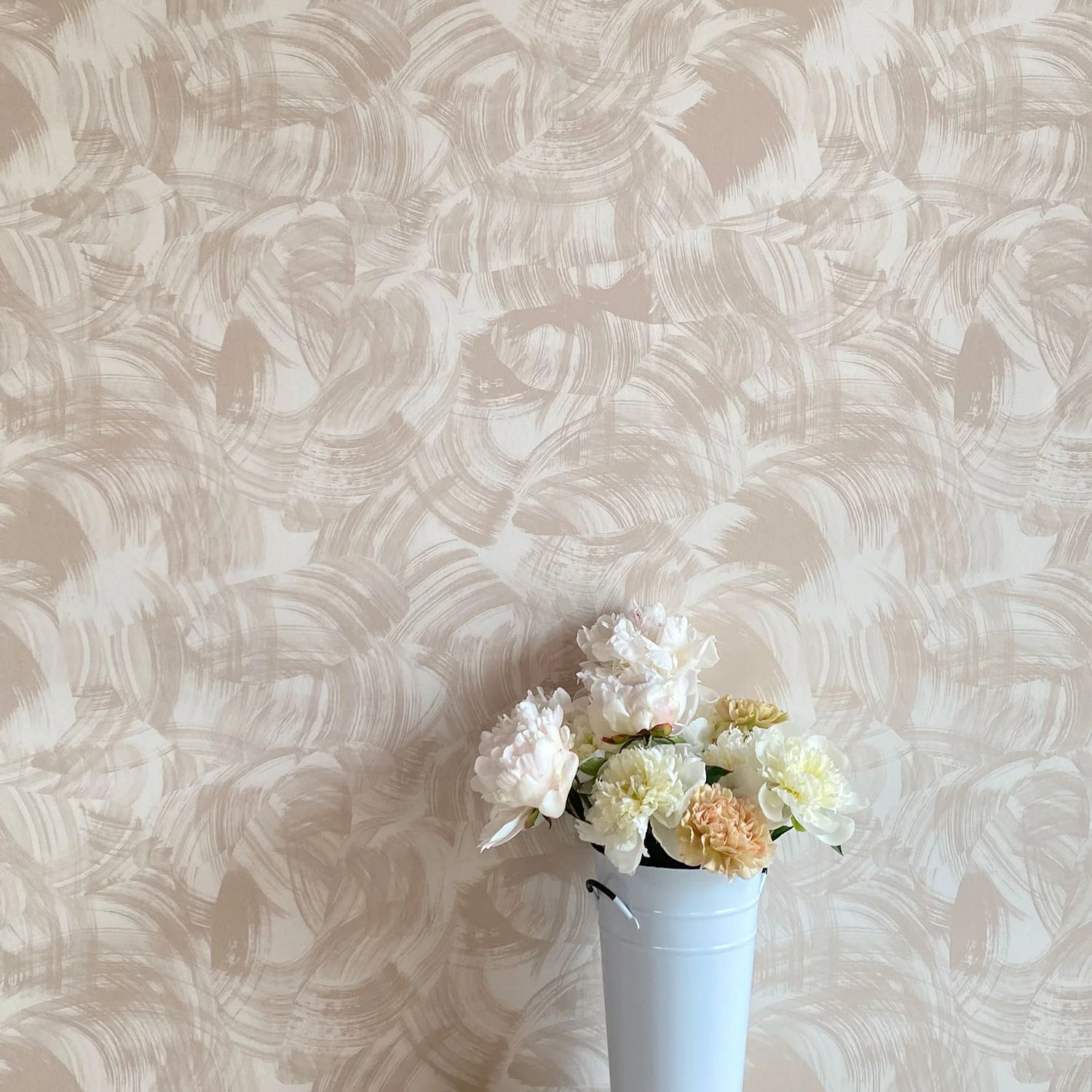 A vase of flowers stands in front of a wall papered in an abstract paint swatch print in tan on a cream field.