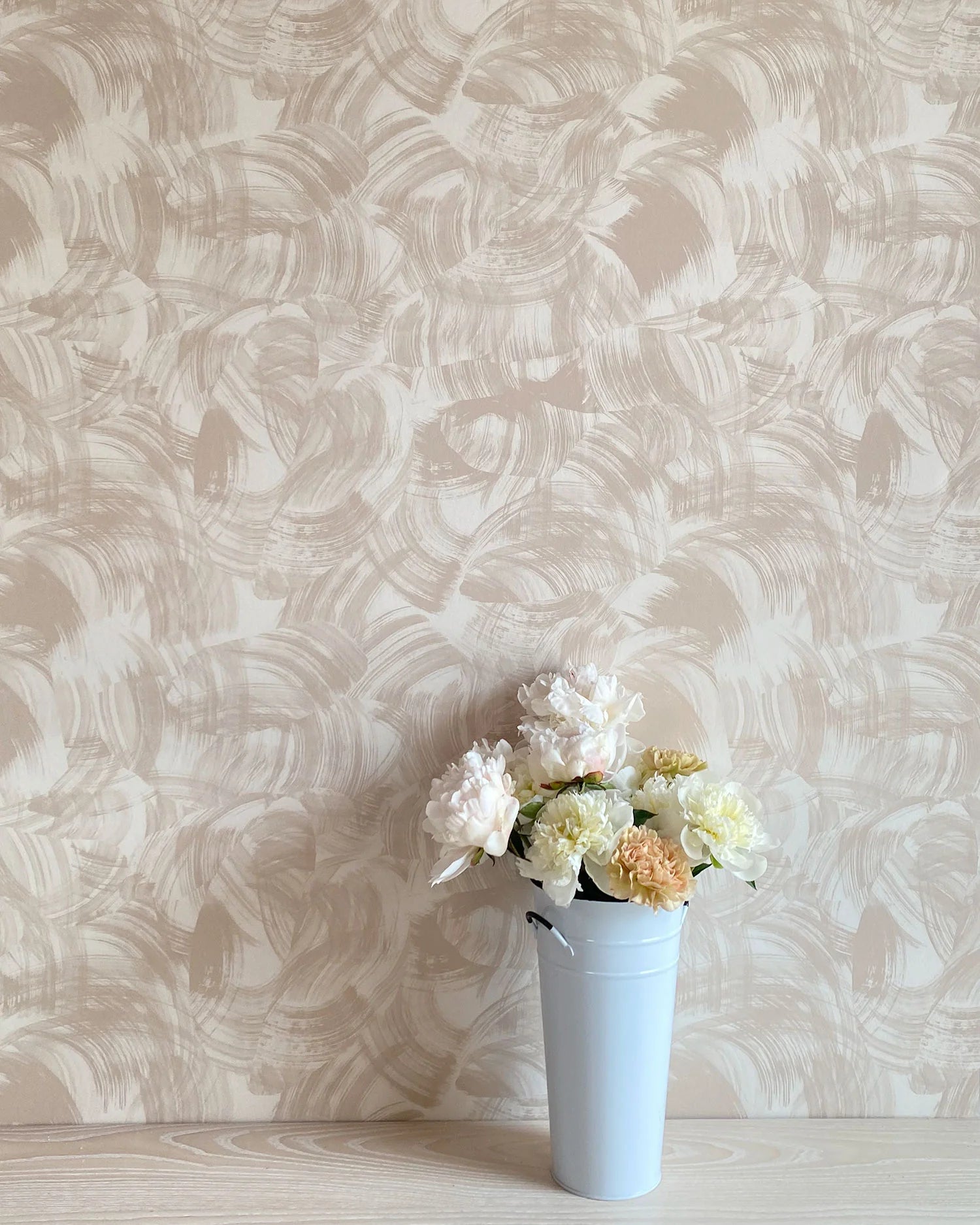 A vase of flowers stands in front of a wall papered in an abstract paint swatch print in tan on a cream field.