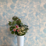 A vase of flowers stands in front of a wall papered in an abstract paint swatch print in light blue on a tan field.