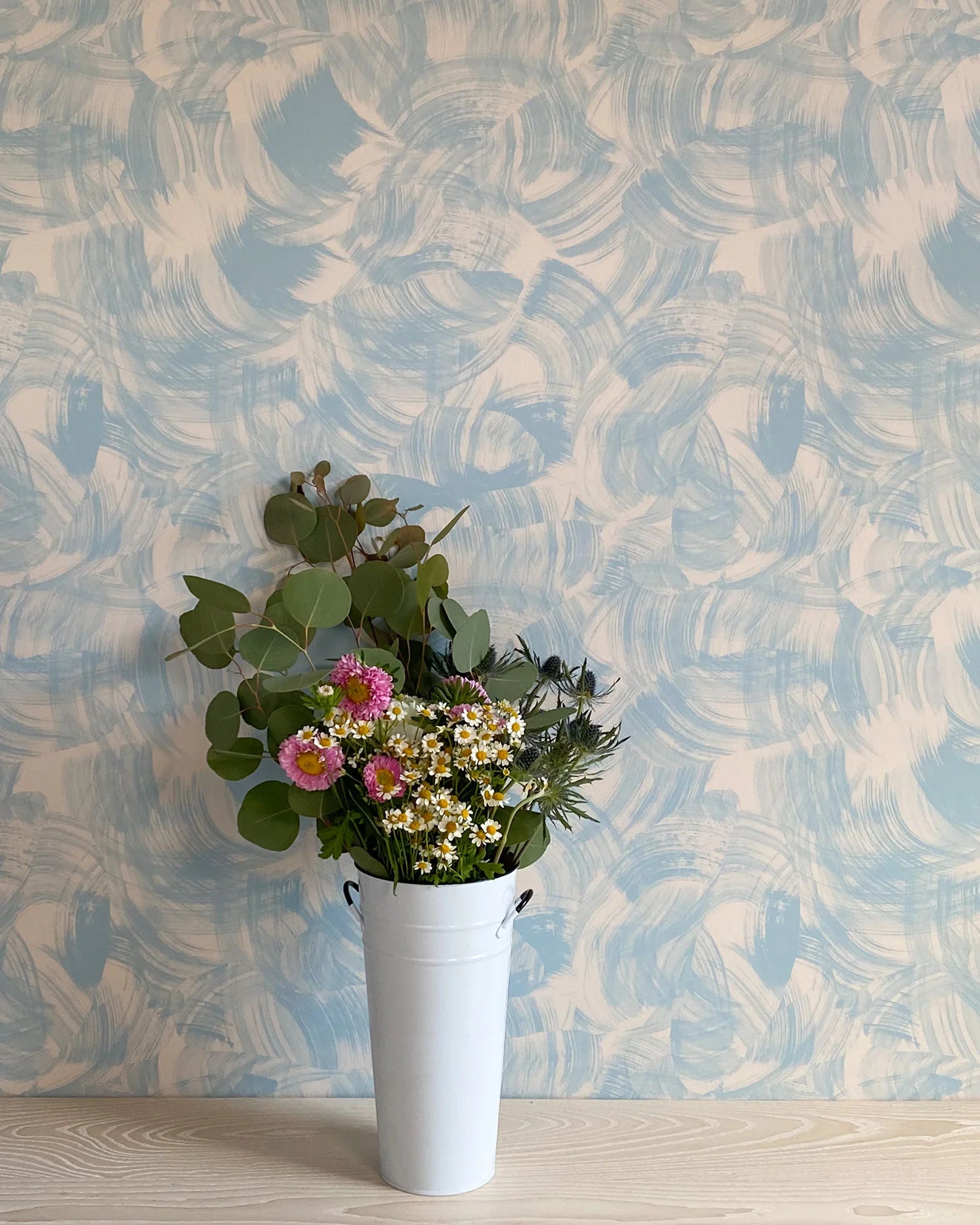 A vase of flowers stands in front of a wall papered in an abstract paint swatch print in light blue on a tan field.