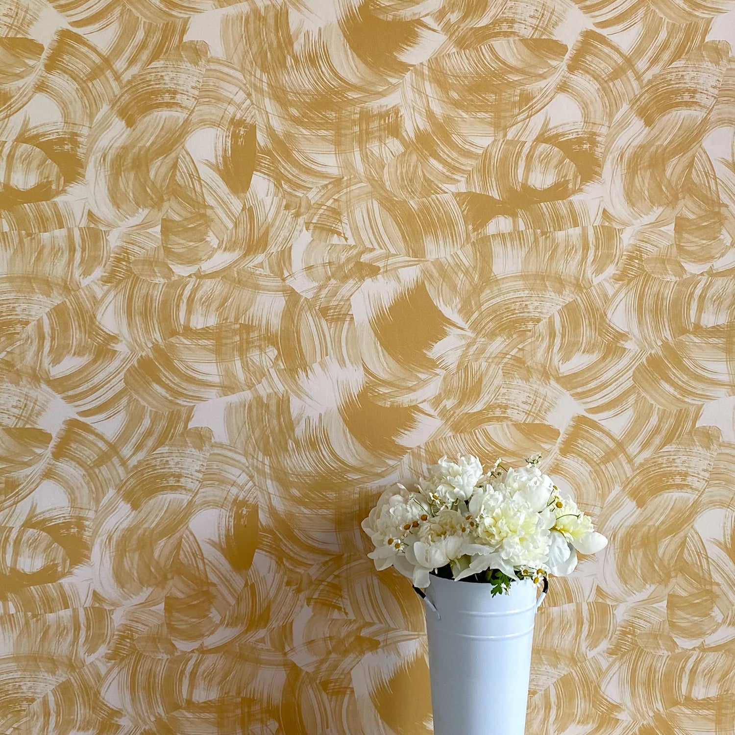 A vase of flowers stands in front of a wall papered in an abstract paint swatch print in mustard on a cream field.