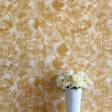 A vase of flowers stands in front of a wall papered in an abstract paint swatch print in mustard on a cream field.