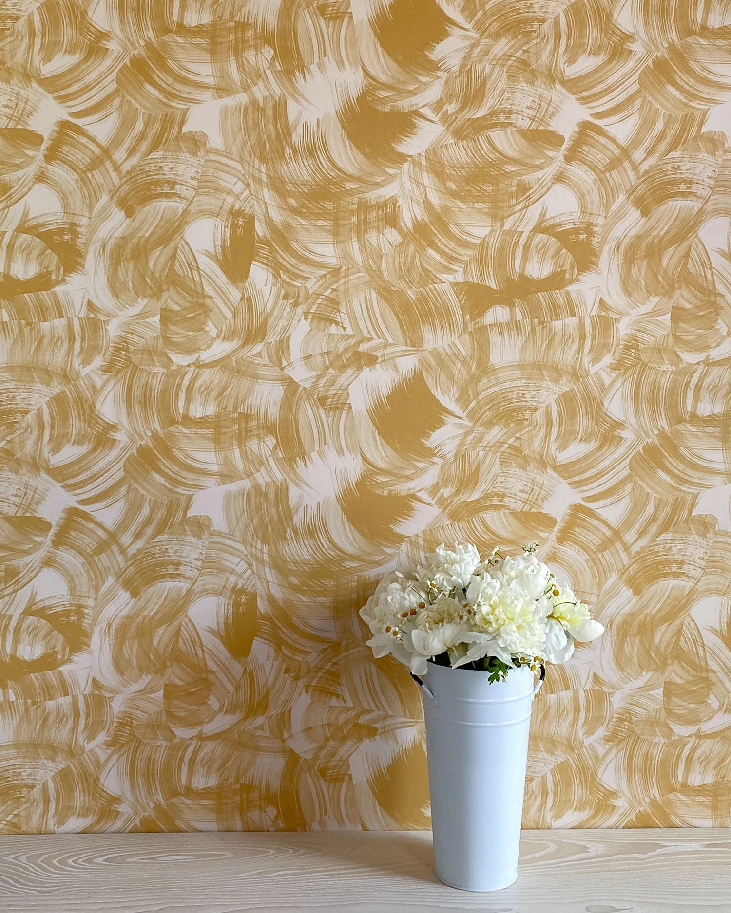 A vase of flowers stands in front of a wall papered in an abstract paint swatch print in mustard on a cream field.