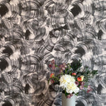 A vase of flowers stands in front of a wall papered in an abstract paint swatch print in black on a white field.