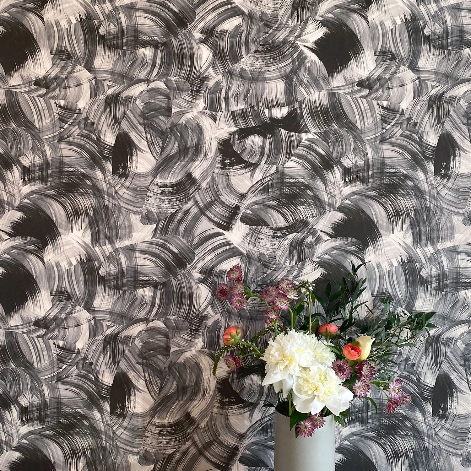 A vase of flowers stands in front of a wall papered in an abstract paint swatch print in black on a white field.