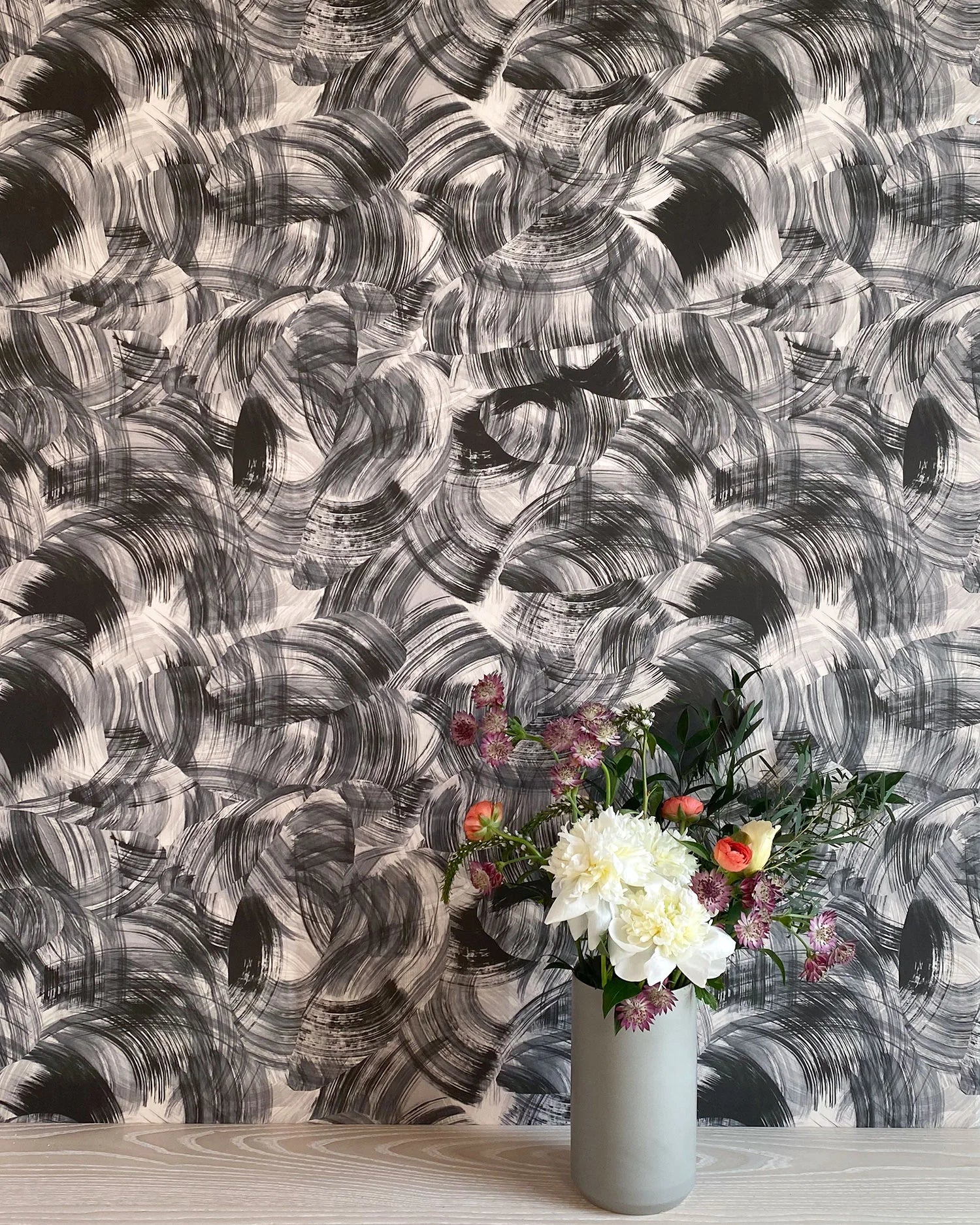 A vase of flowers stands in front of a wall papered in an abstract paint swatch print in black on a white field.