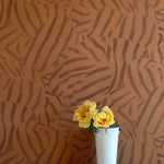 A vase of flowers stands in front of a wall papered in a playful broken stripe print in dark brown on a brown field.