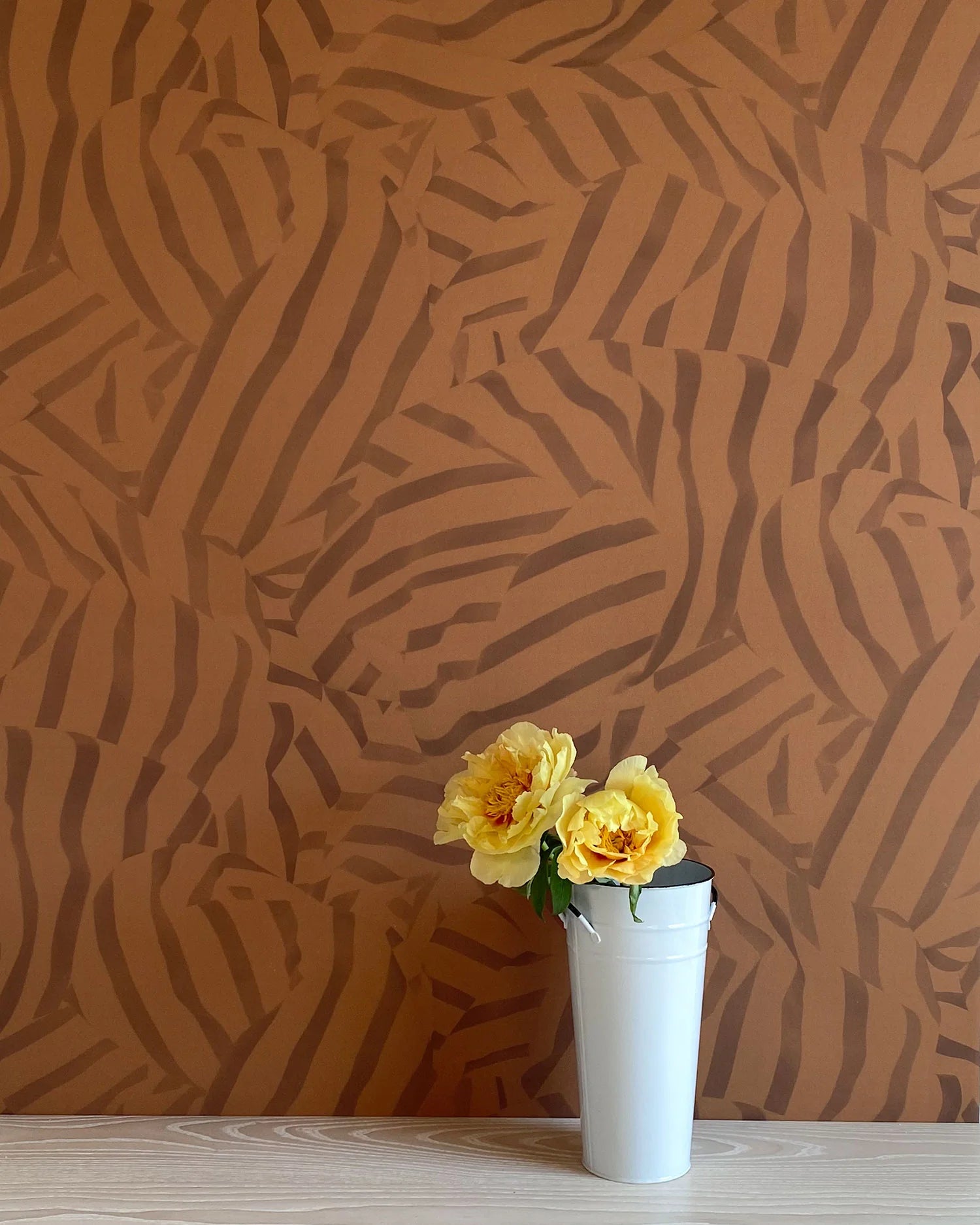 A vase of flowers stands in front of a wall papered in a playful broken stripe print in dark brown on a brown field.