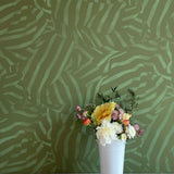 A vase of flowers stands in front of a wall papered in a playful broken stripe print in dark green on a green field.