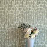 A vase of flowers stands in front of a wall papered in a linear floral print in blue and white on a green field.