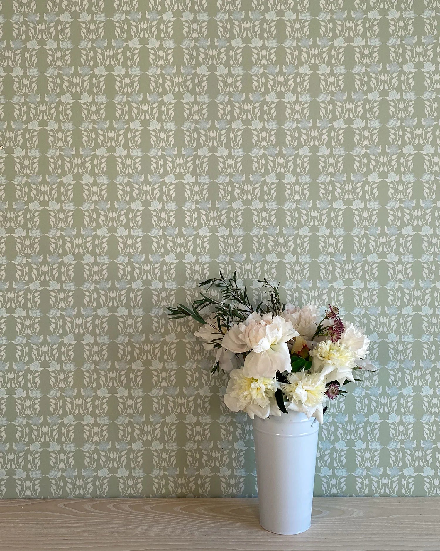 A vase of flowers stands in front of a wall papered in a linear floral print in blue and white on a green field.