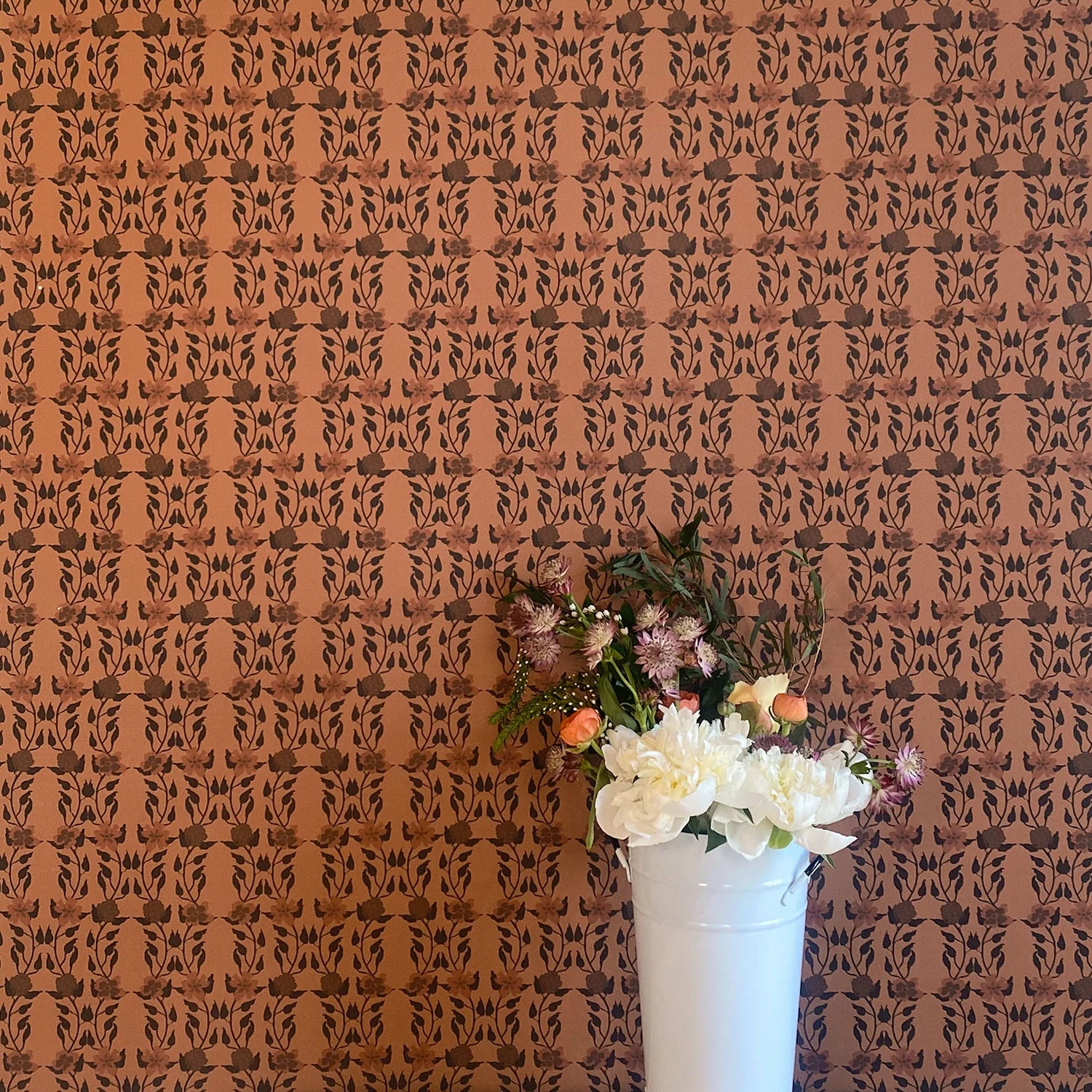 A vase of flowers stands in front of a wall papered in a linear floral print in red and black on an orange field.