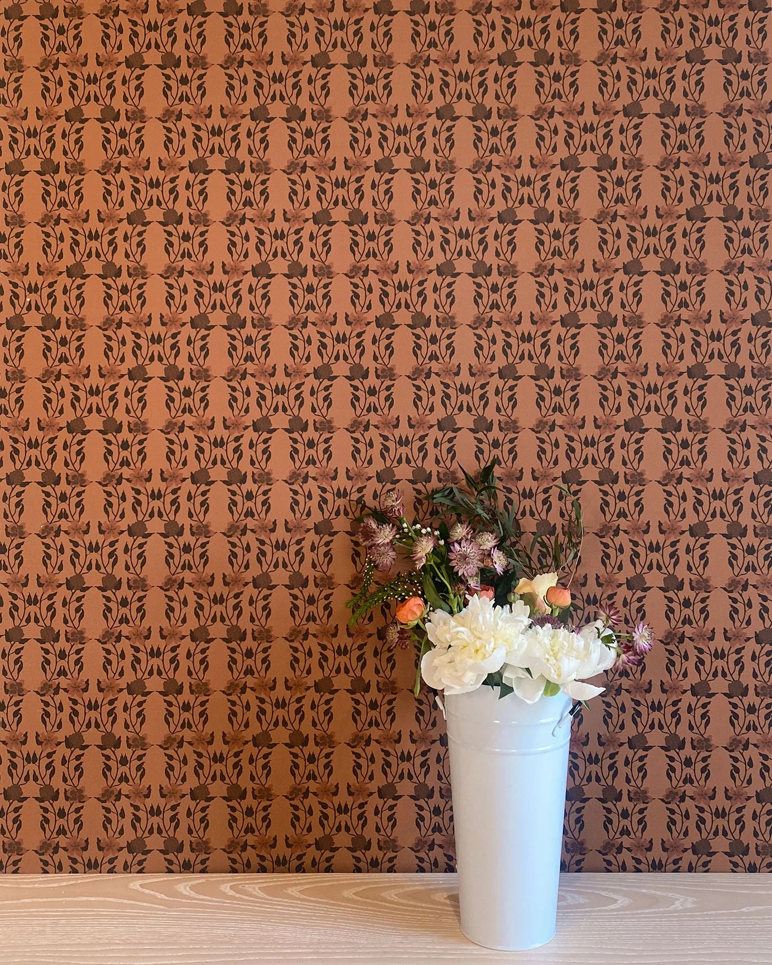 A vase of flowers stands in front of a wall papered in a linear floral print in red and black on an orange field.