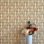 A vase of flowers stands in front of a wall papered in a linear floral print in red, orange and green on a cream field.