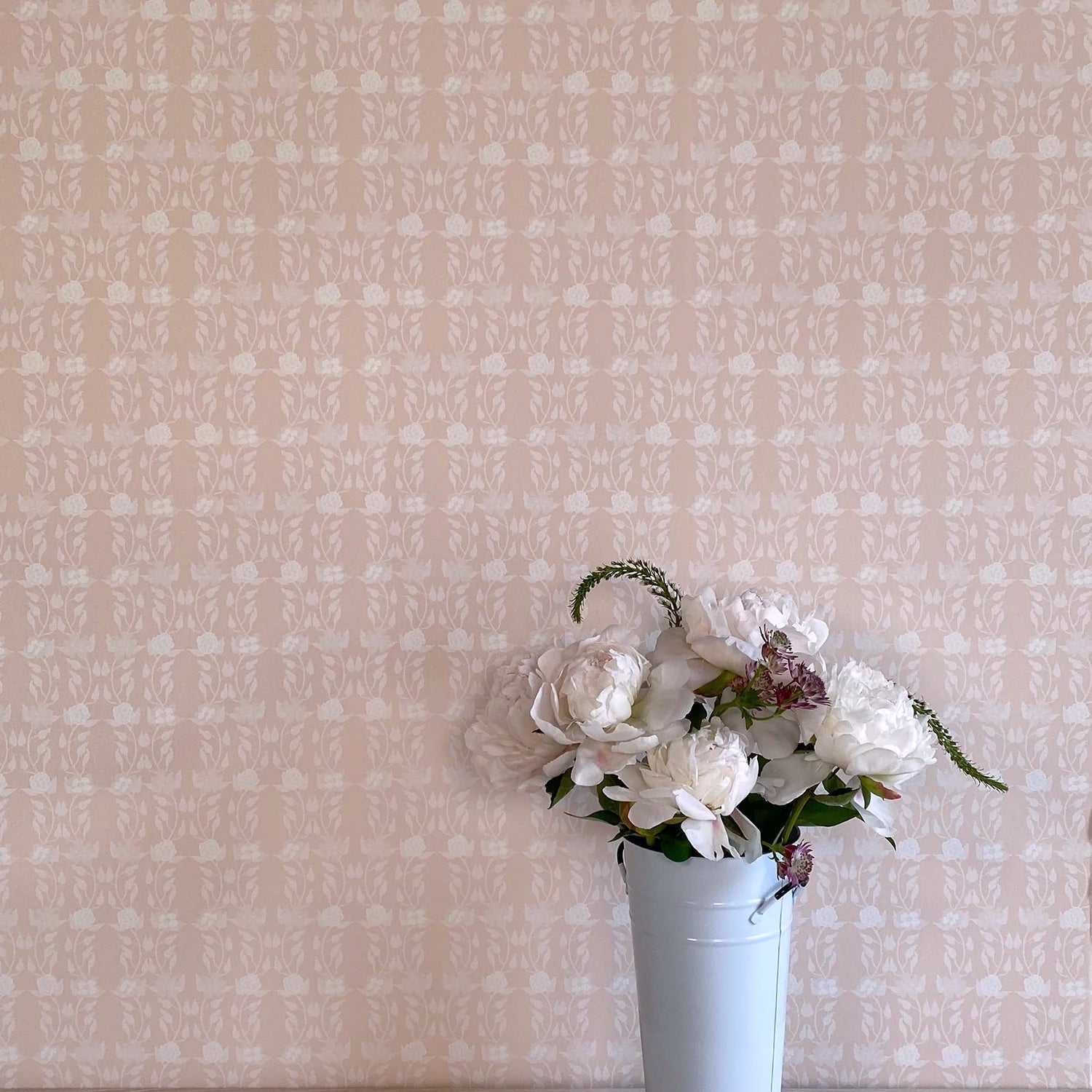 A vase of flowers stands in front of a wall papered in a linear floral print in white and cream on a light pink field.