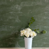 A vase of flowers stands in front of a wall papered in an abstract textural pattern in shades of green.