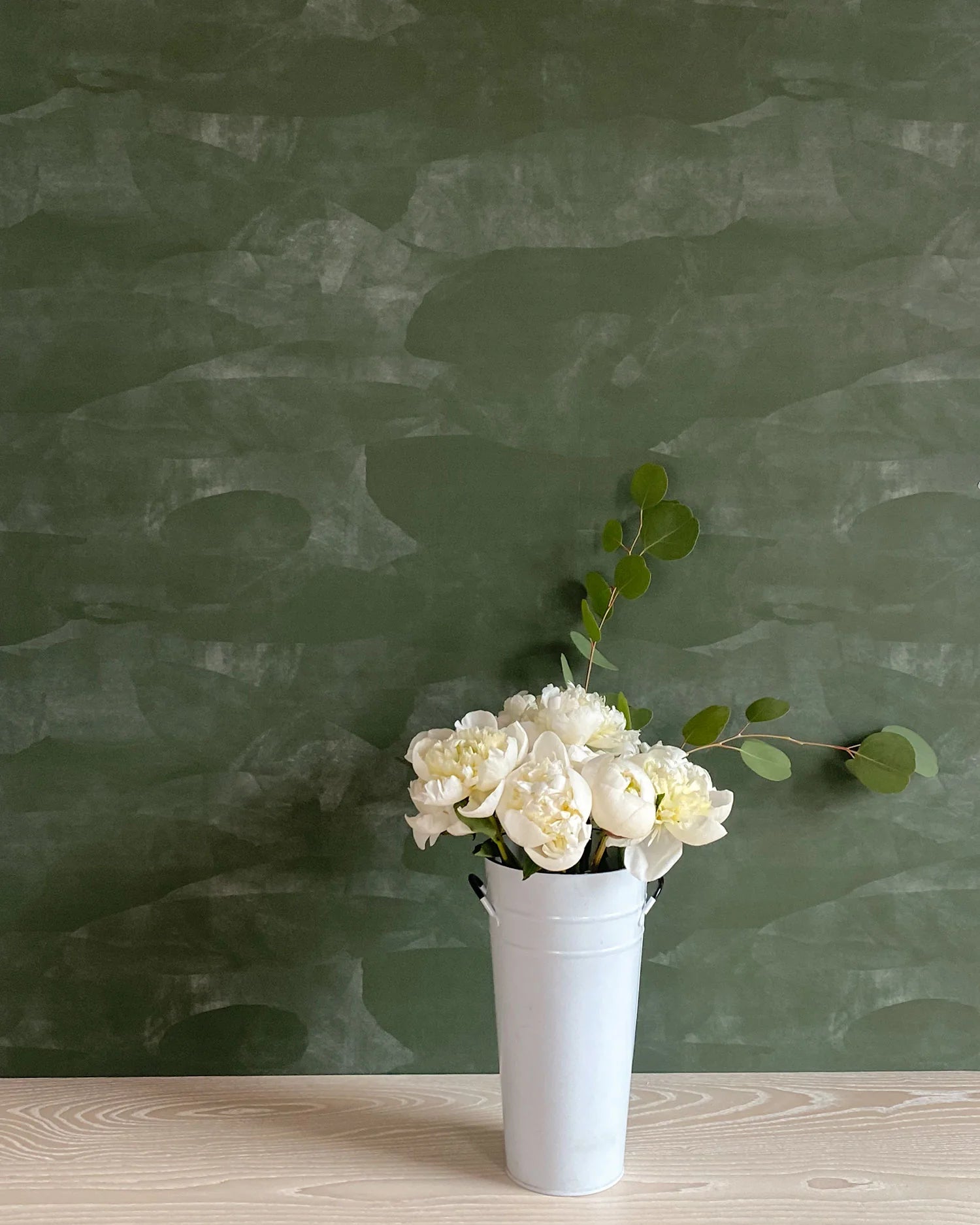 A vase of flowers stands in front of a wall papered in an abstract textural pattern in shades of green.